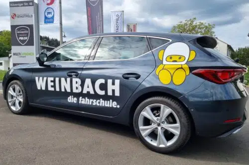 Dunkelgrauer PKW mit weißer Beschriftung der Fahrschule Weinbach und gelbem Maskottchen