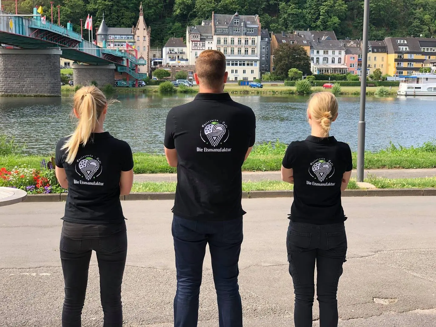 Drei Mitarbeitende in schwarzen Poloshirts mit bedrucktem Logo am Moselufer in Traben-Trarbach