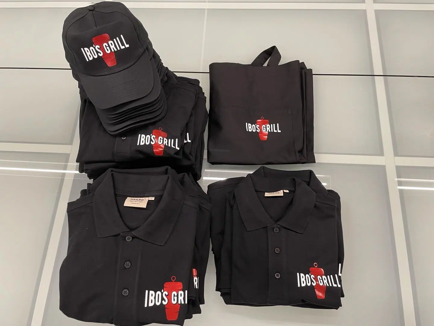Schwarze Poloshirts, Caps und Schürzen mit bedrucktem Logo Ibo's Grill